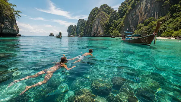 Travellers snorkeling in crystal-clear turquoise water near tropical limestone islands and longtail boats during a Phuket island experience in southern Thailand.